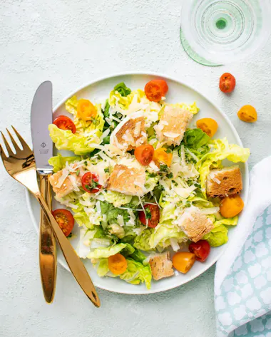 Salată fresh cu salată verde, roșii cherry, crutoane și brânză, servită pe un platou alb cu tacâmuri aurii.