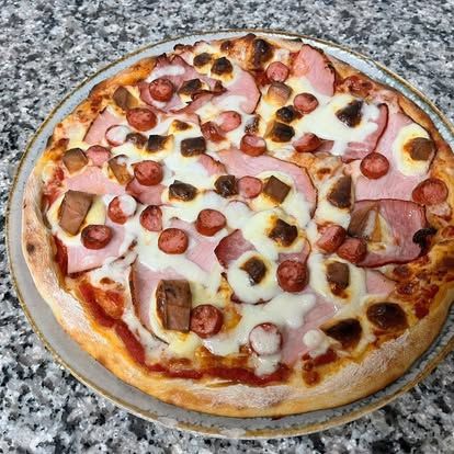 Pizza cu șuncă, cârnați și brânză, servită pe un platou de piatră, cu margine crocantă.
