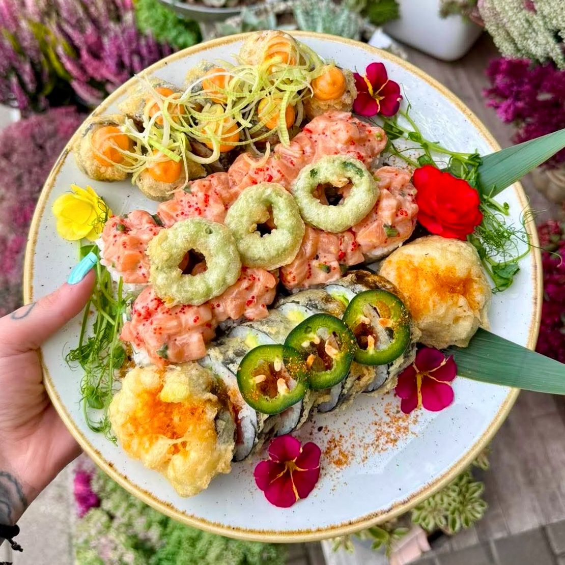 Talerz sushi z różnorodnymi rolkami, dekoracjami kwiatowymi i ziołami, w otoczeniu roślin.