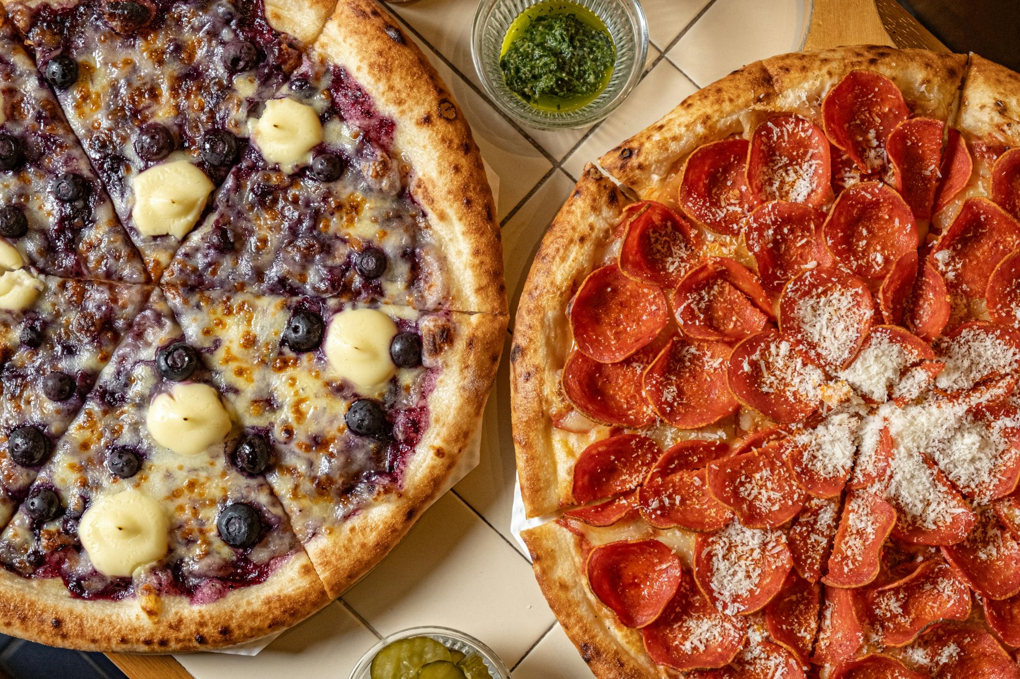 Dwie pizze: jedna z serem i jagodami, druga z pepperoni i parmezanem, podane z sosami.