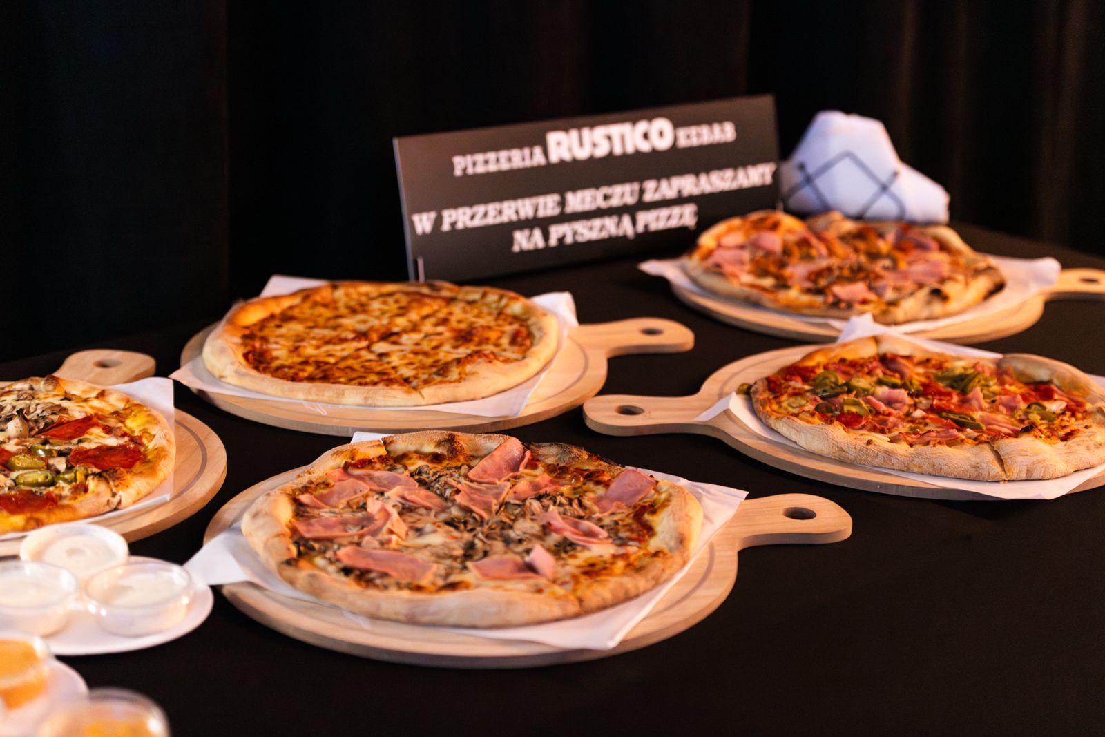 Cztery rodzaje pizzy na drewnianych łapkach, w tle napis "Pizzeria Rustico", podana w eleganckim otoczeniu.