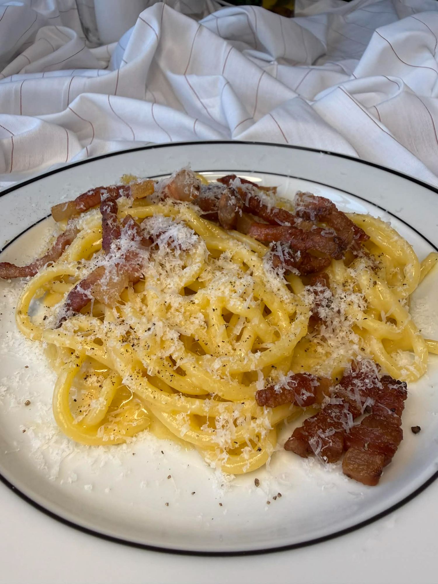 Makaron carbonara z boczkiem, serem i przyprawami, podany na talerzu w białym obrusie.