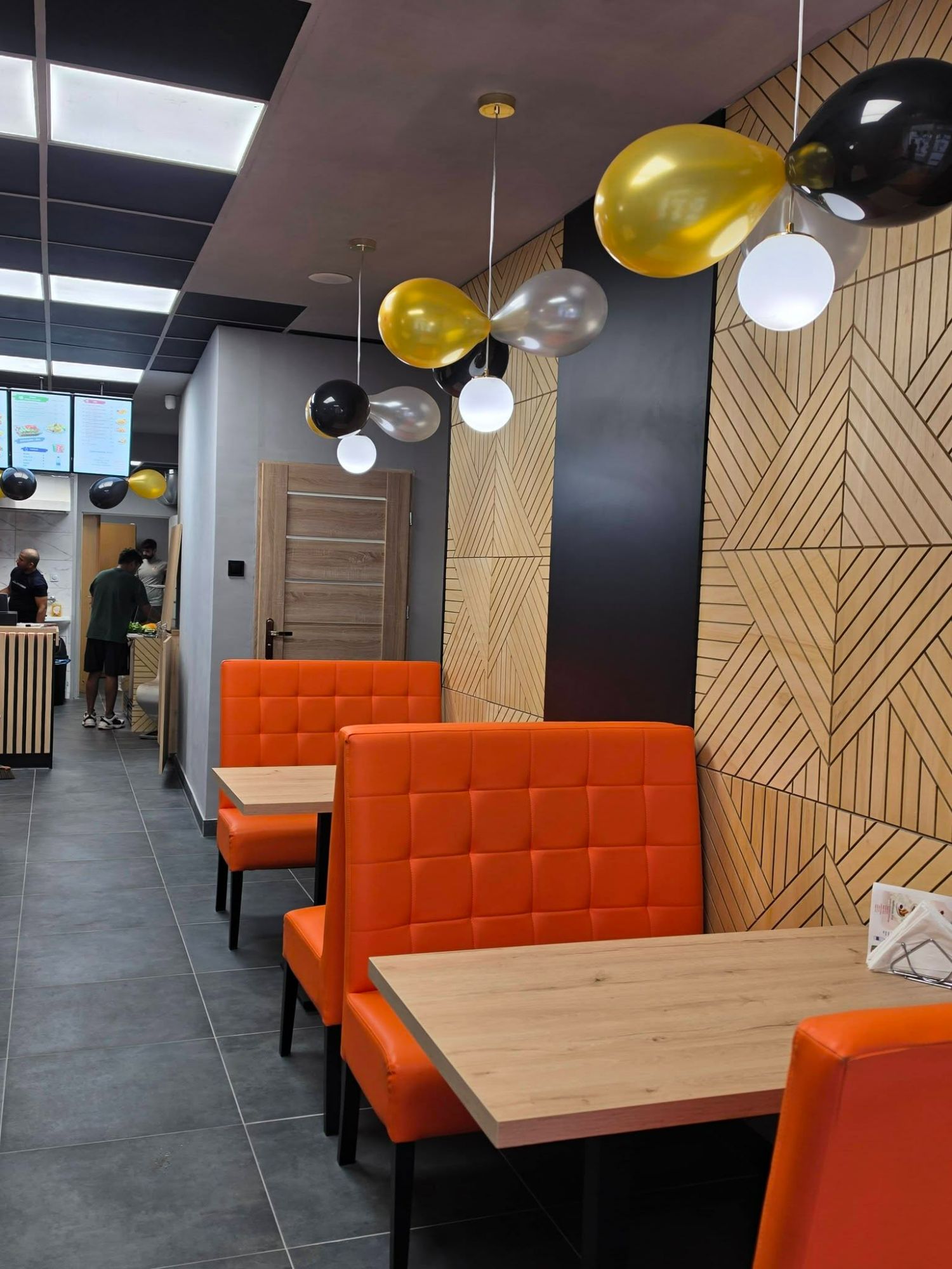 Nowoczesna wnętrze restauracji z pomarańczowymi siedzeniami, drewnianymi detalami i balonami w kolorach czarnym, złotym i białym.