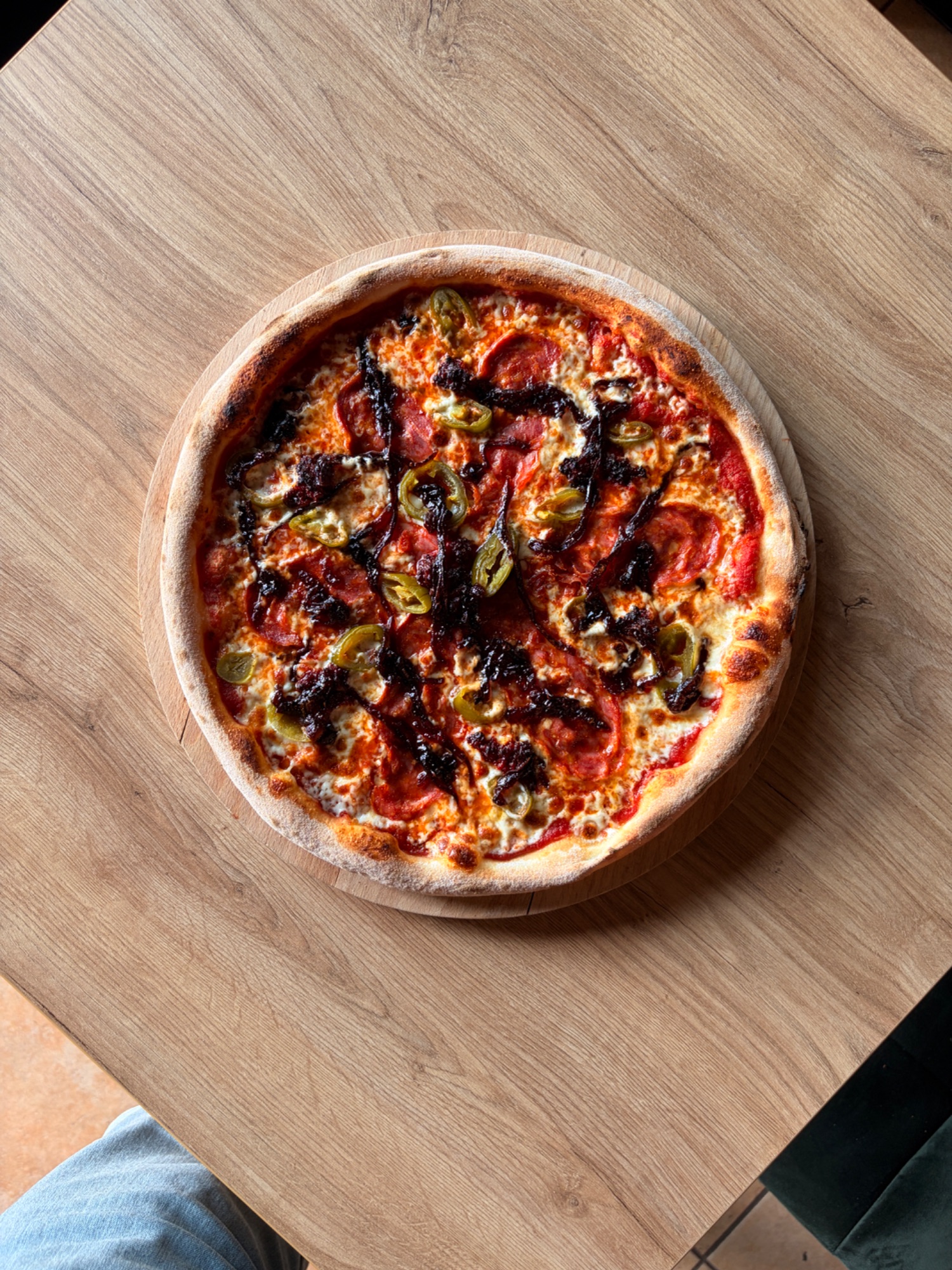 Pizza na drewnianej desce, z sosem pomidorowym, serem, kiełbasą i papryczkami, na tle drewnianego stołu.