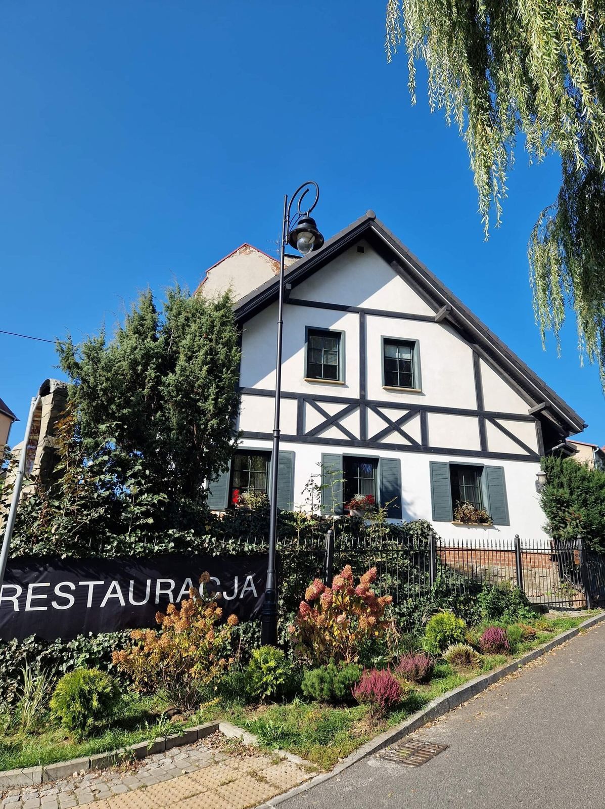 Biały budynek z drewnianymi detalami, ogrodzony, z napisem "Restauracja" i zielenią wokół.