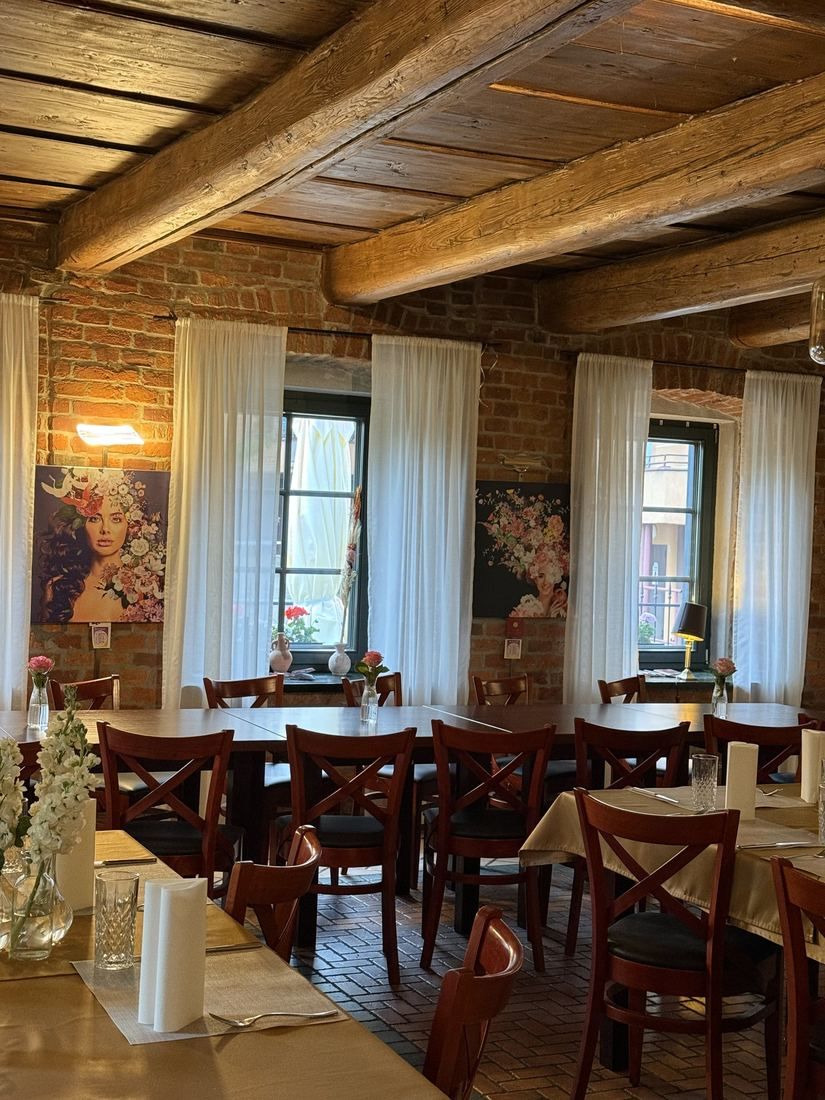 Wnętrze restauracji z drewnianymi belkami, oknami, białymi firankami i eleganckimi stołami.