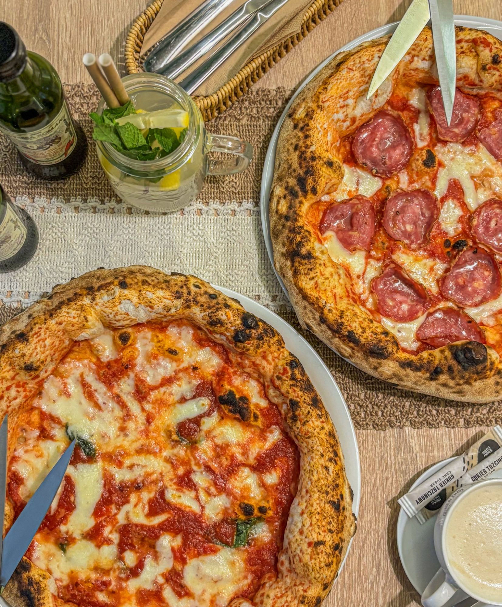 Dwa rodzaje pizzy na stole, jeden z serem i pomidorami, drugi z salami, obok napój cytrynowy w szklance.