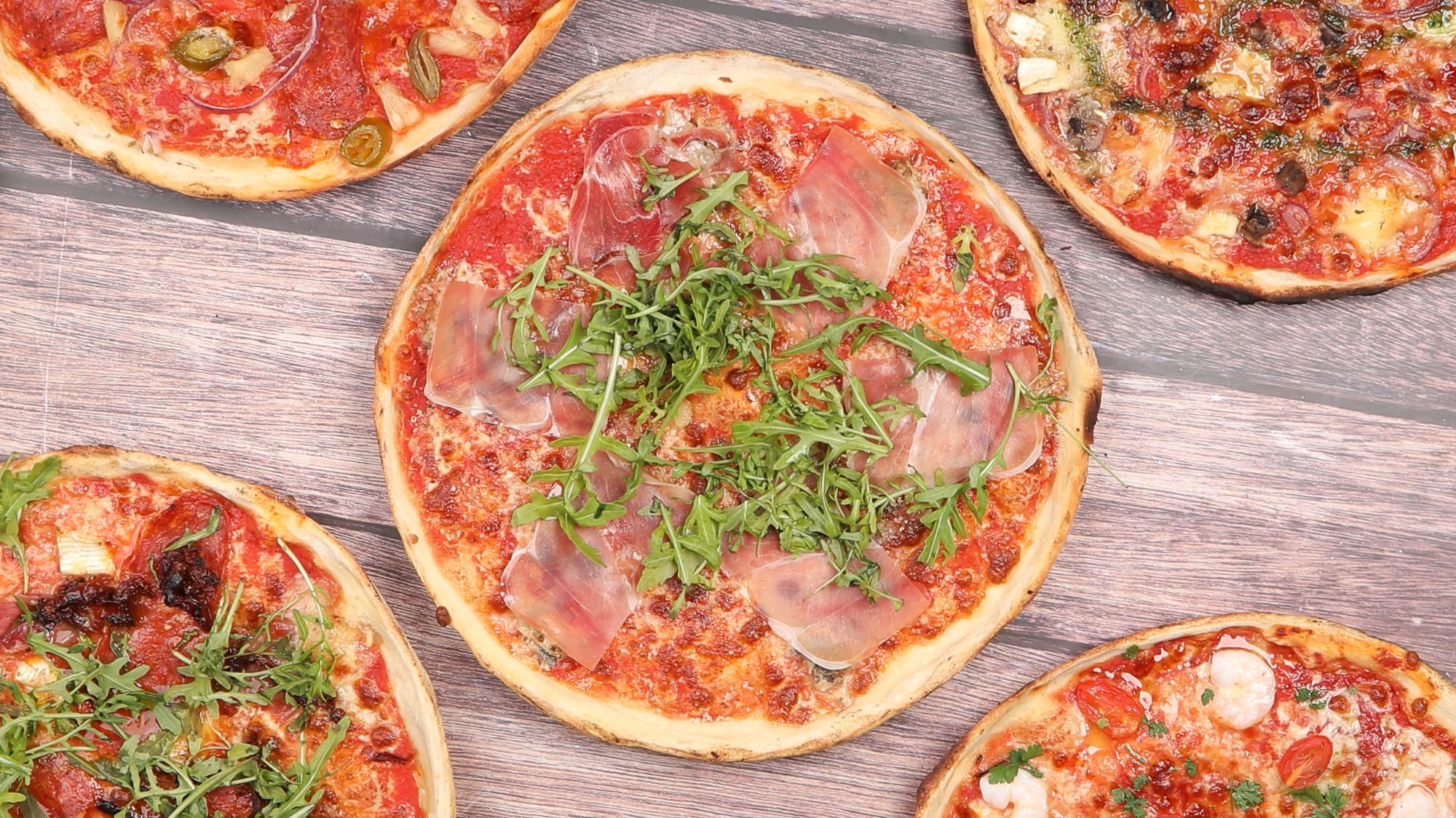 Różne pizze na drewnianym stole, w tym jedna z szynką i rukolą, obok innych smaków.