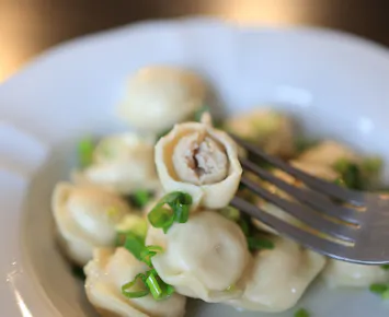 Pierogi z mięsem, posypane posiekaną cebulką, na białym talerzu z widelcem w środku.