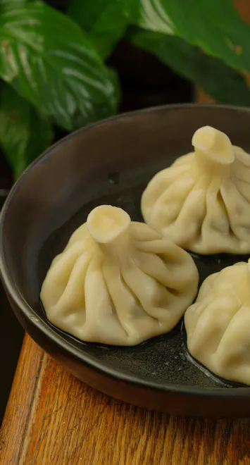 Trzy pierożki na talerzu, przypominające sakiewki, z liściem rośliny w tle.
