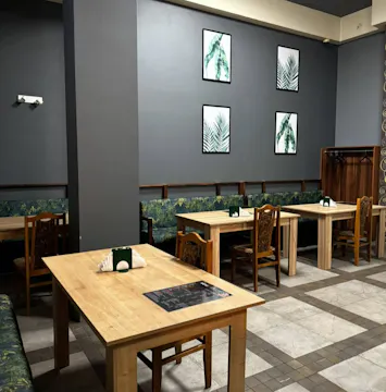 Wnętrze restauracji z drewnianymi stołami, krzesłami i zielonymi obrazami na szarych ścianach.