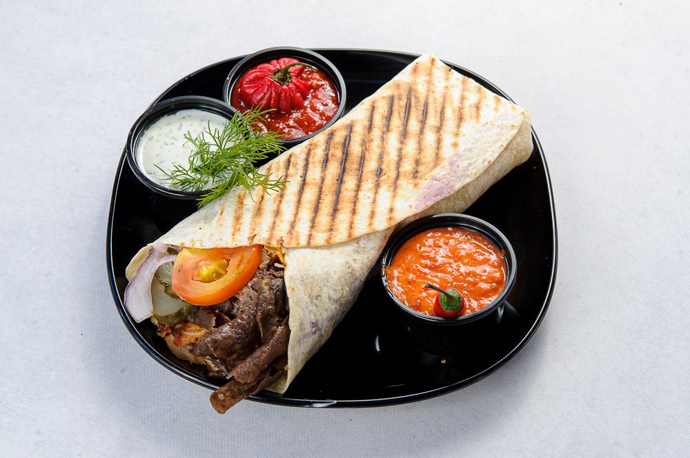 Tortilla z wołowiną, pomidorem, sałatą oraz sosem, podana na czarnym talerzu z dodatkowymi sosami.