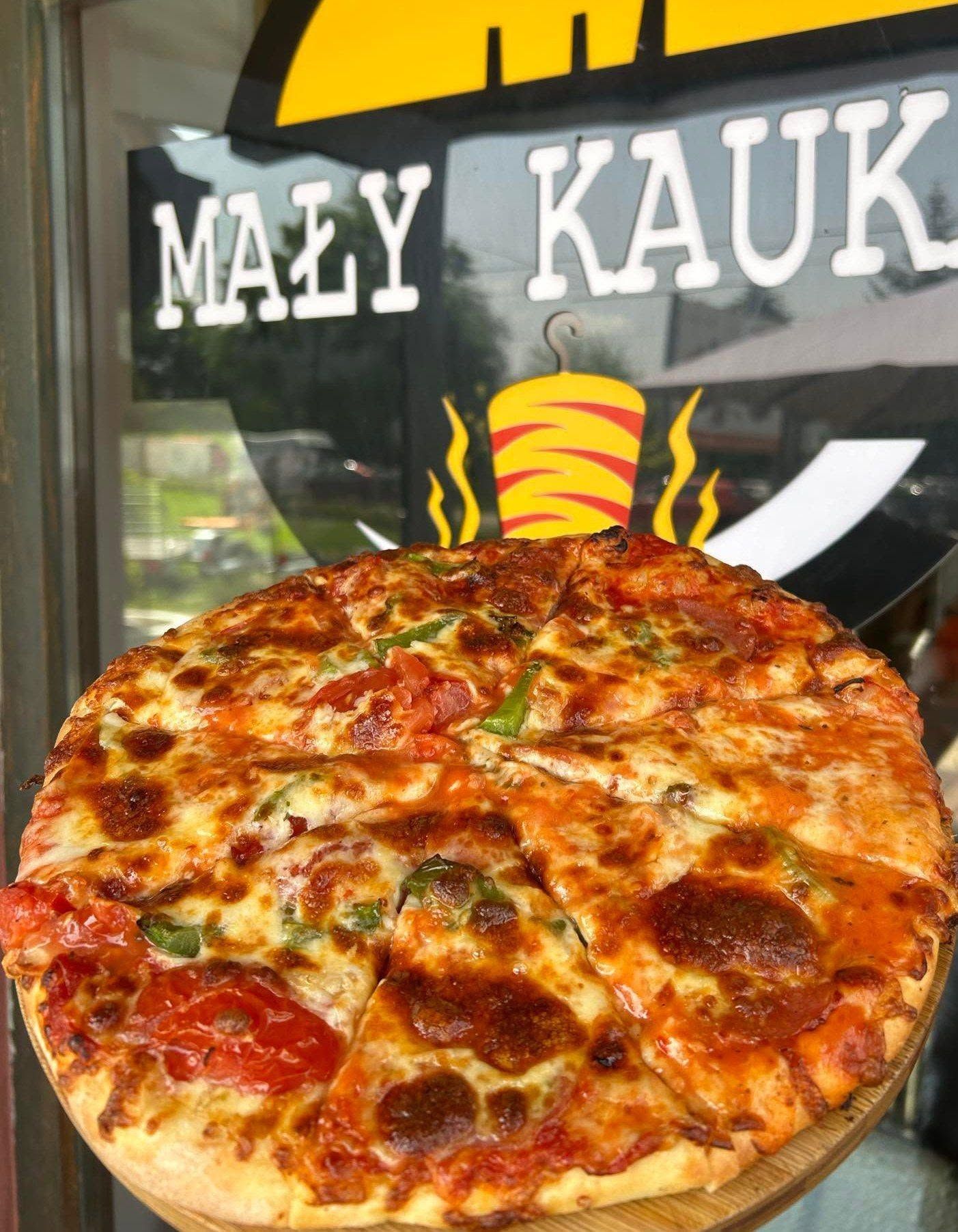 Pizza z serem, pomidorami i papryką na drewnianej desce, z logo restauracji "Mały KauK" w tle.