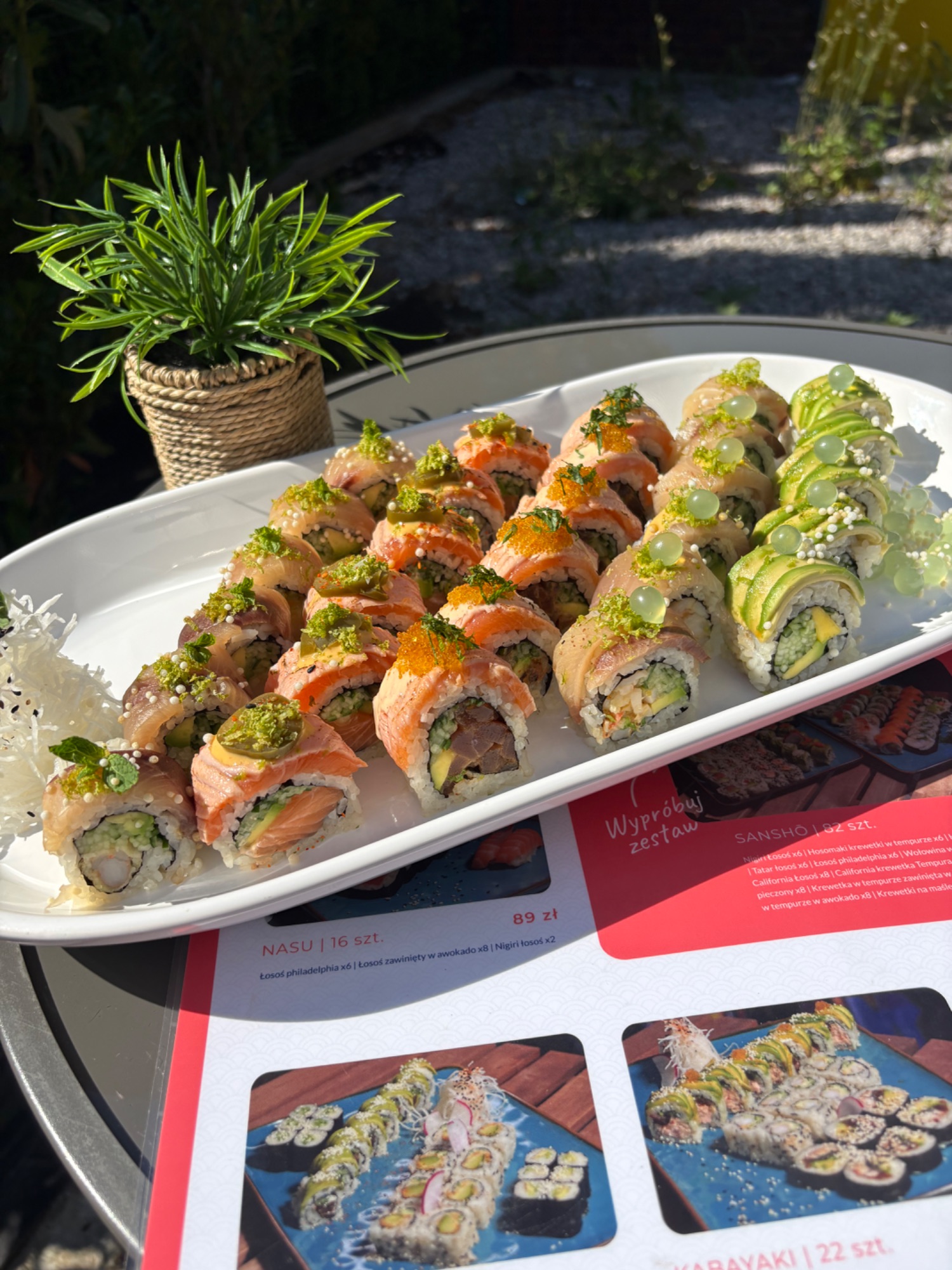 Talerz z różnorodnym sushi, w tym rolki z rybą, warzywami i sezamem, na tle zieleni.