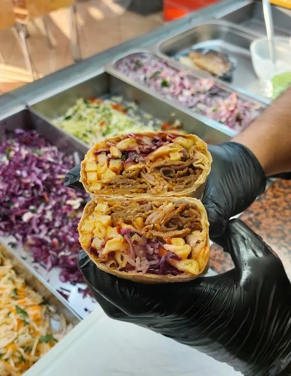 Podzielony tortilla z mięsem i warzywami, trzymany w rękawicy, w tle kolorowe składniki.