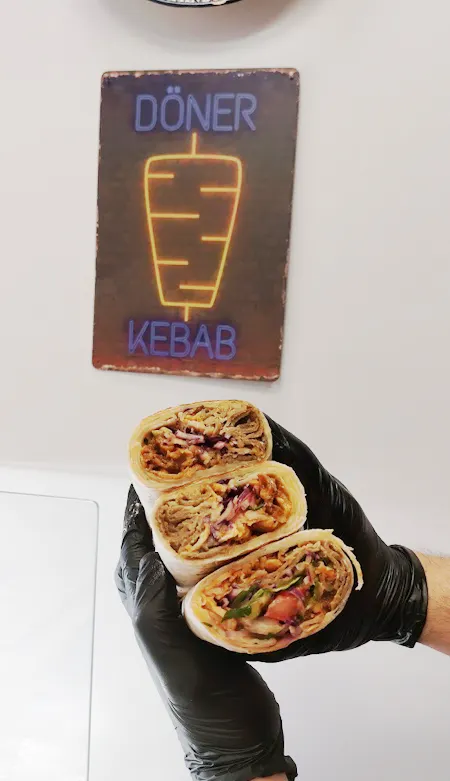 Ręka trzymająca trzy kebaby w tortilli, w tle neonowy znak "Döner Kebab".