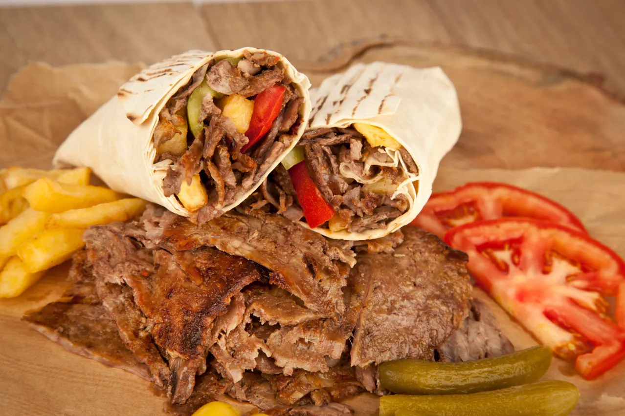 Burrito z mięsem, warzywami i frytkami, podane na drewnianym talerzu obok świeżych plastrów pomidora i ogórka.