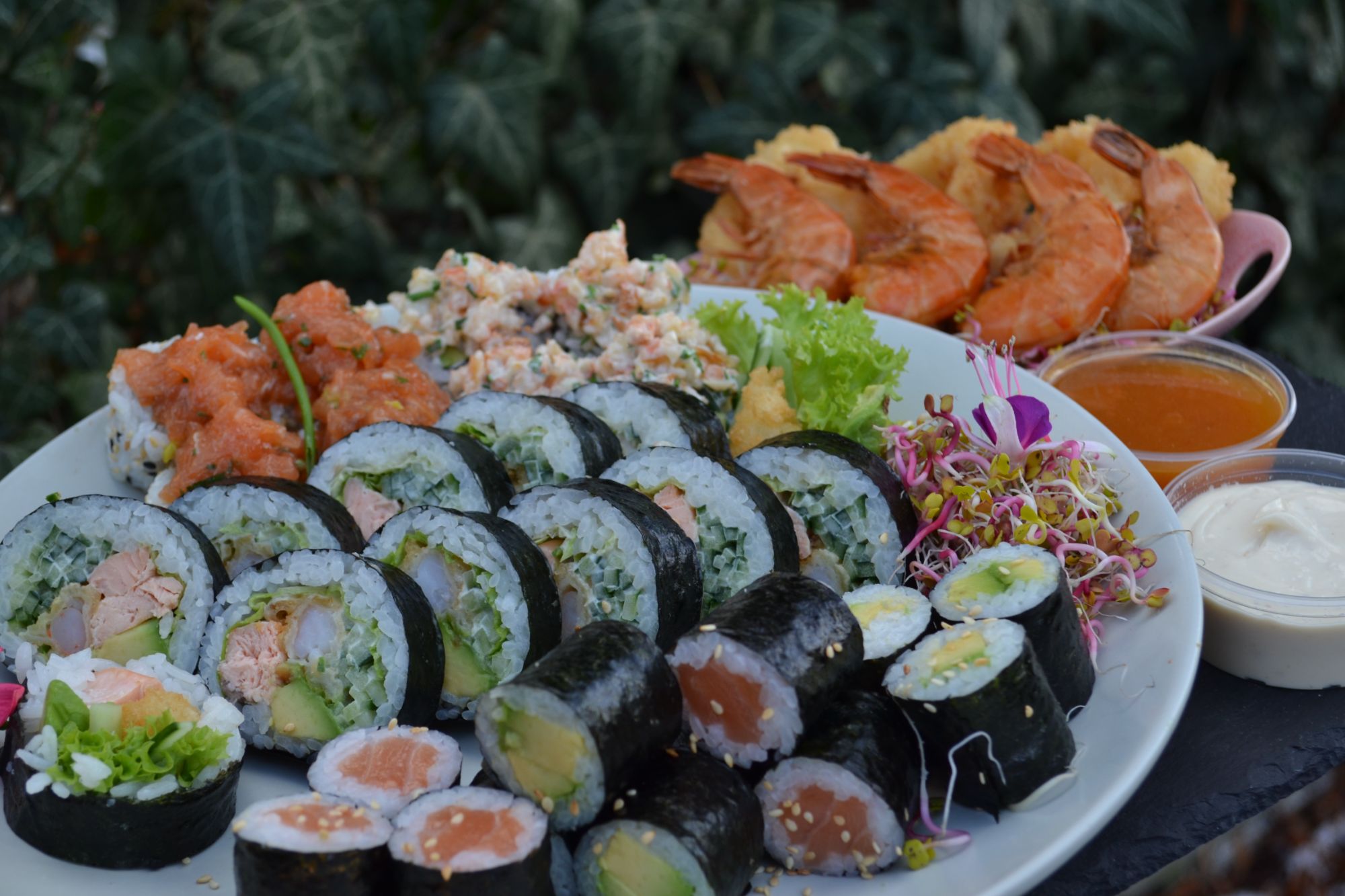 Talerz sushi z rolkami, krewetkami, sałatką i dodatkami na tle zieleni.