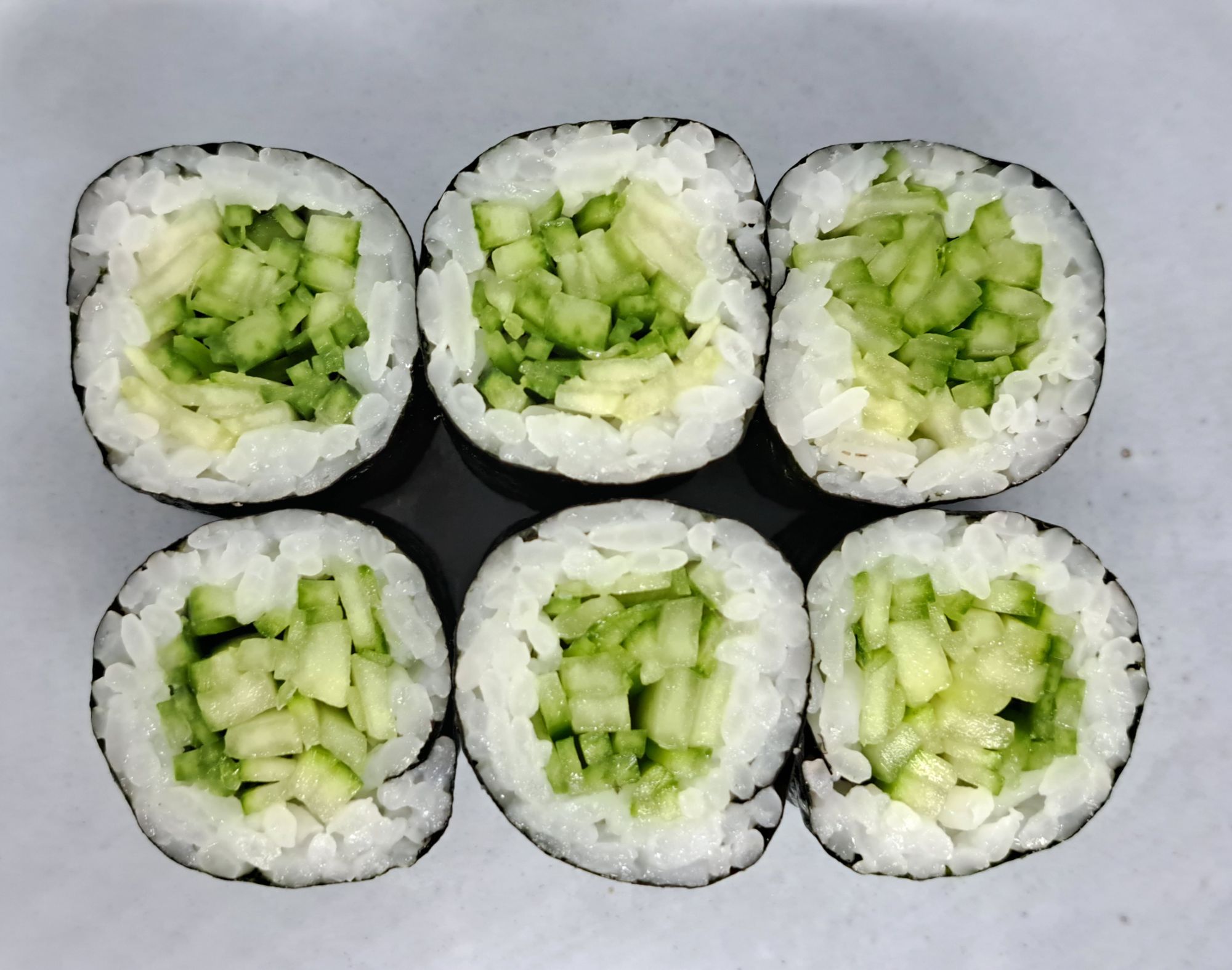 Sześć rolek sushi z ryżem, ogórkiem i awokado, ułożonych w rządzie na szarym talerzu.