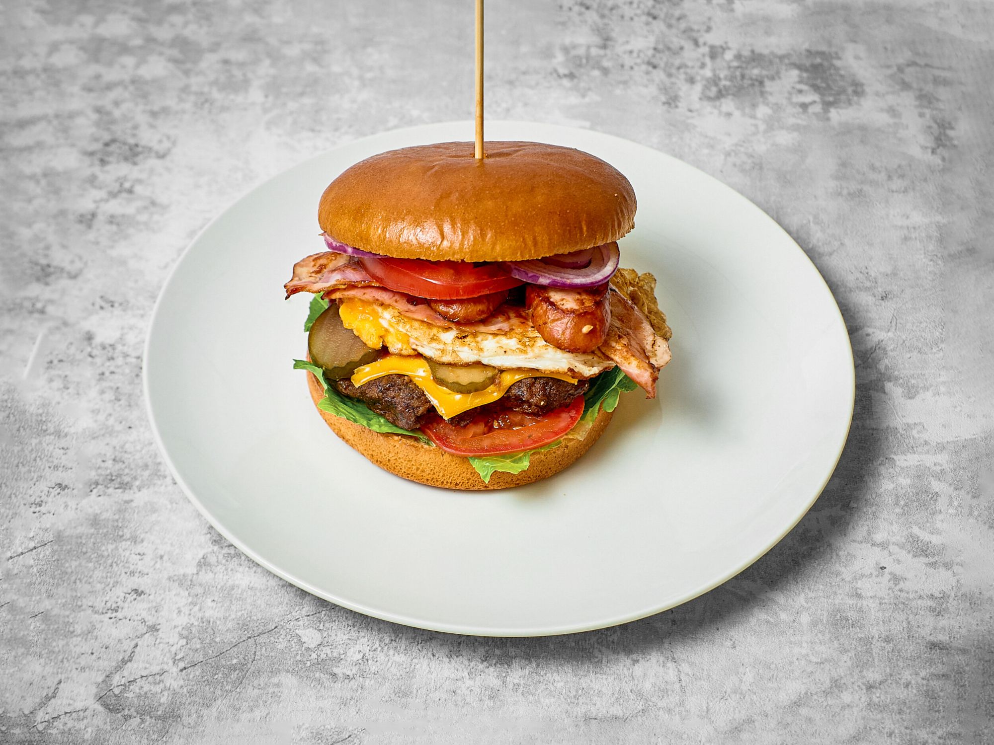 Burger z wołowiną, serem cheddar, jajkiem, boczkiem, pomidorem i sałatą na talerzu.
