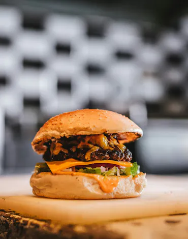 Soczysty hamburger z wołowiną, serem cheddar, sałatą i dodatkami na desce, w tle rozmyta kuchnia.