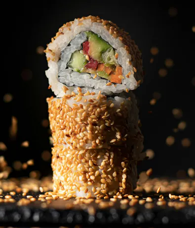 Trzy kawałki sushi z warzywami, obtoczone w sezamie, na tle ciemnego tła z rozpryskującymi się ziarenkami sezamu.