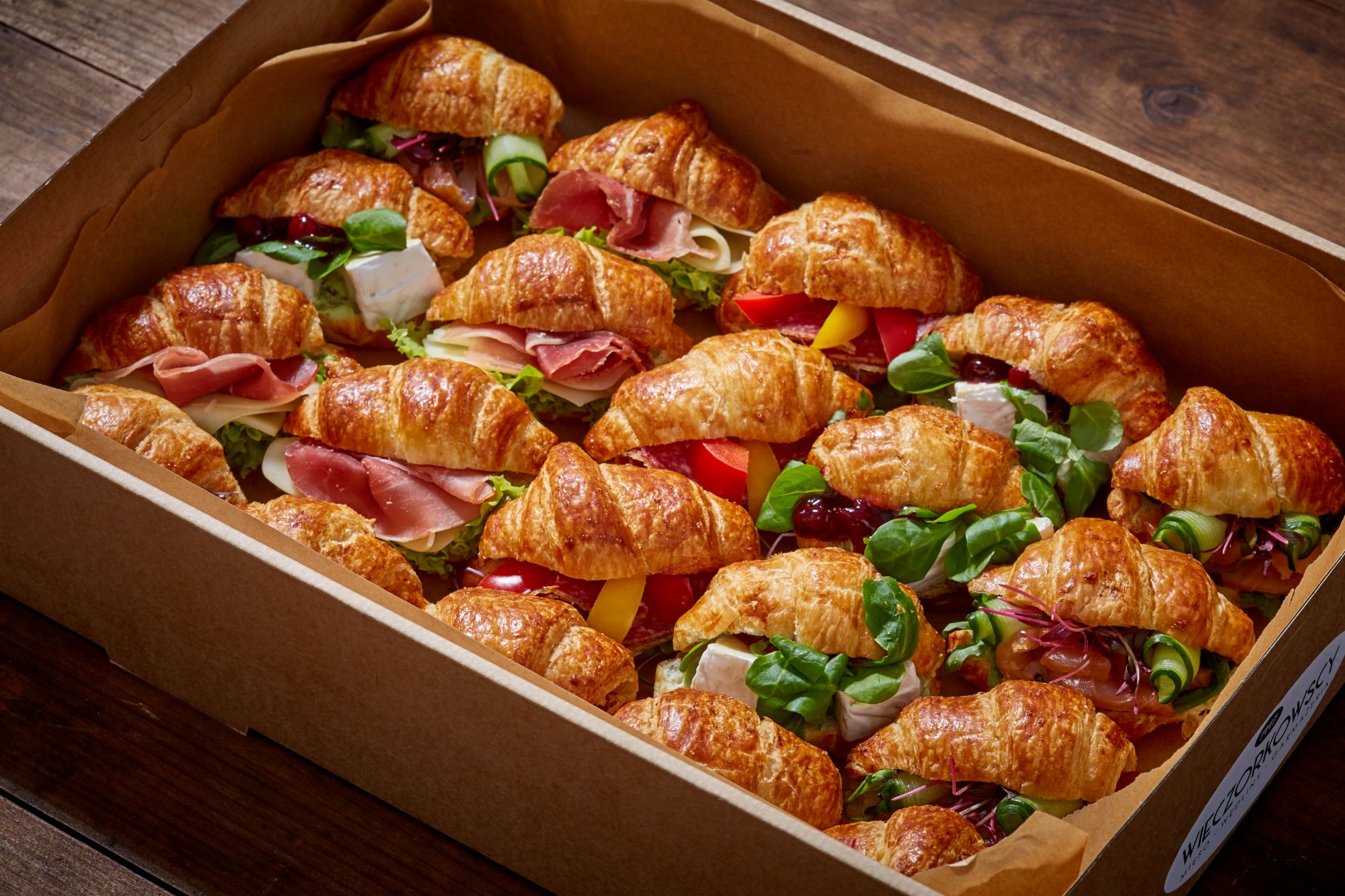 Skrzynka z mini croissantami z szynką, serem, warzywami i dżemem, świeżo przygotowanymi i podanymi na drewnianym tle.