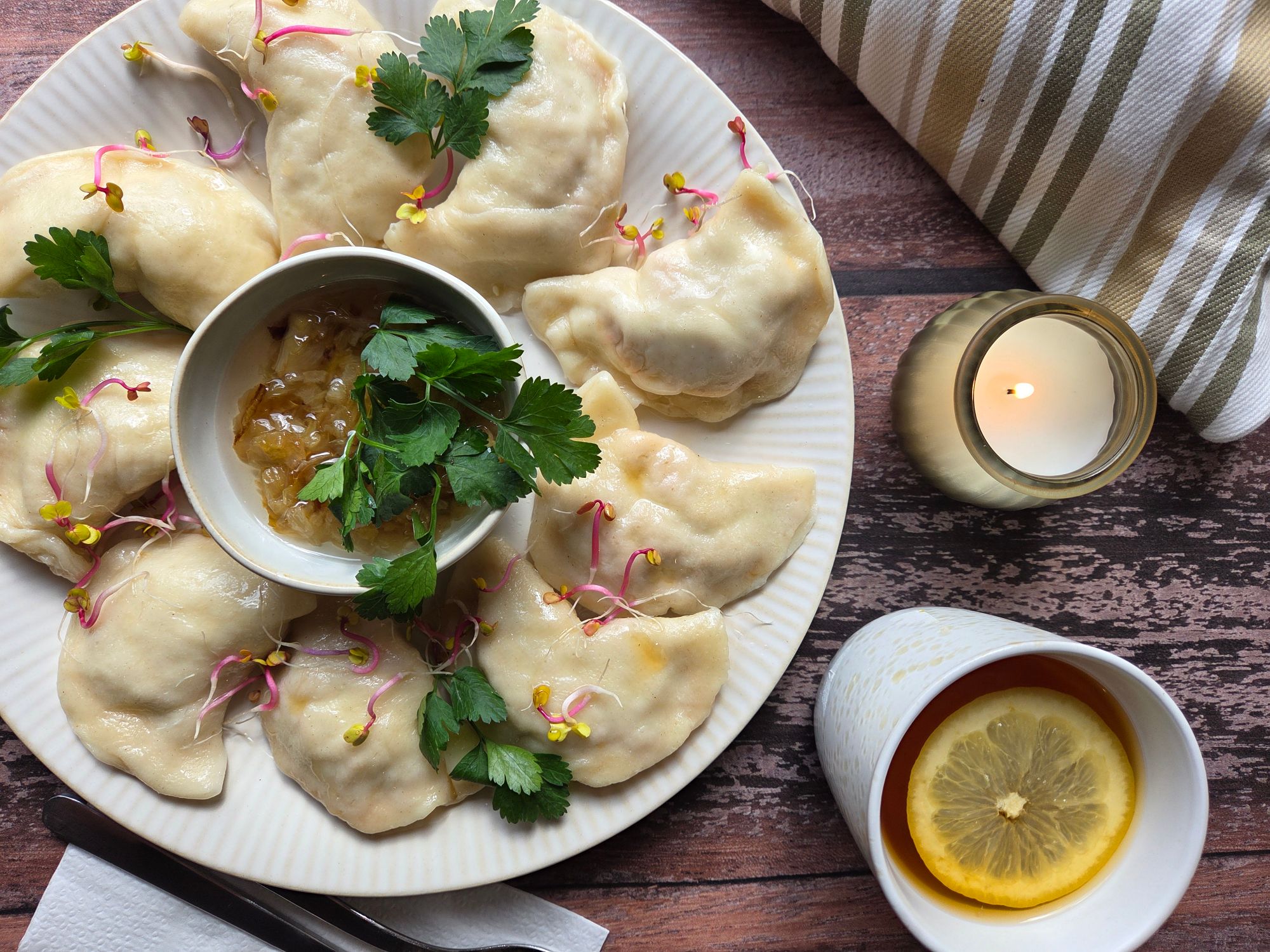 Pierogi na talerzu z sosikiem, świeżą kolendrą i kiełkami, obok świeca i filiżanka z cytryną.