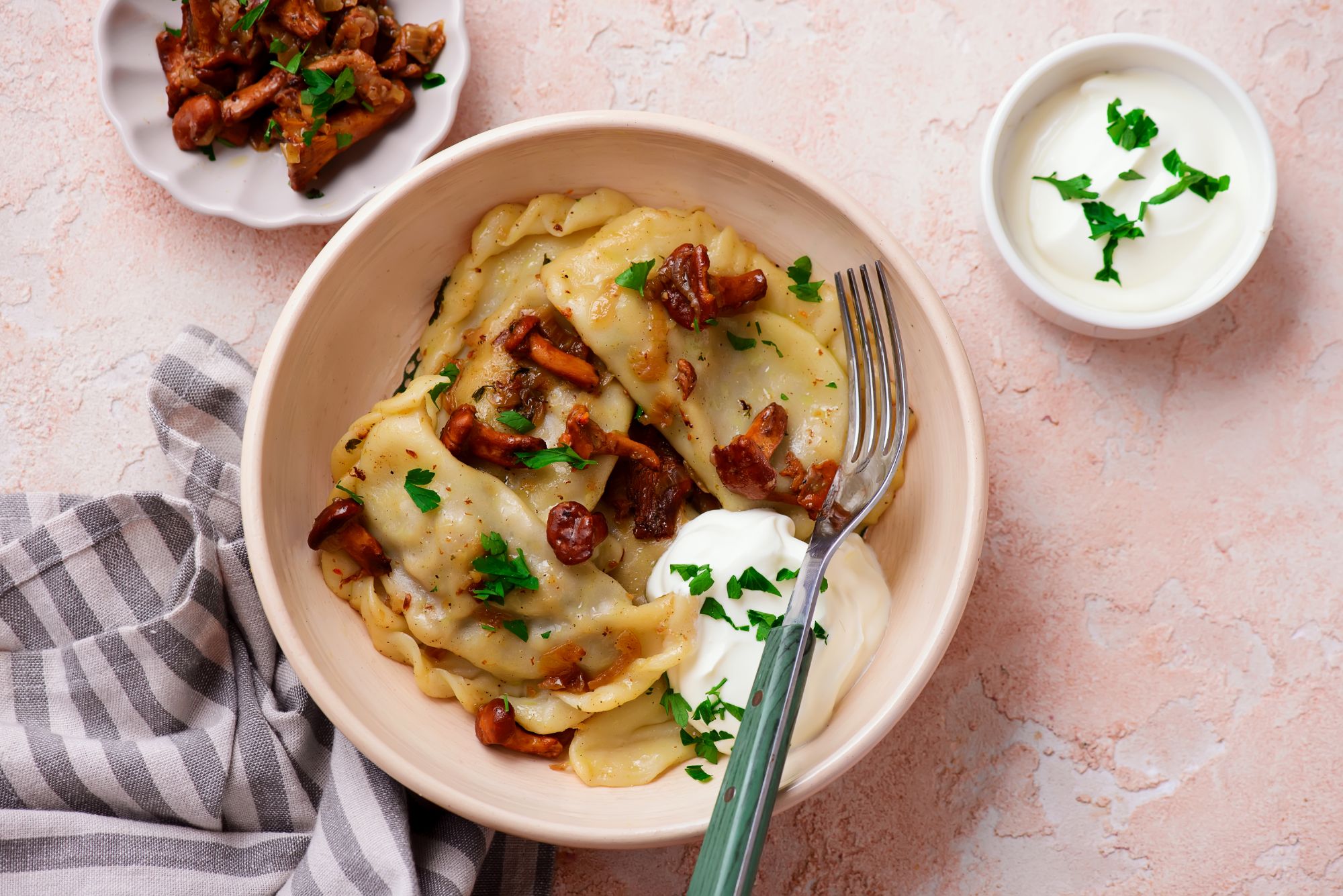 Zastawa talerza z pierogami, śmietaną i dodatkową porcją grzybów na boku, udekorowane świeżą natką pietruszki.