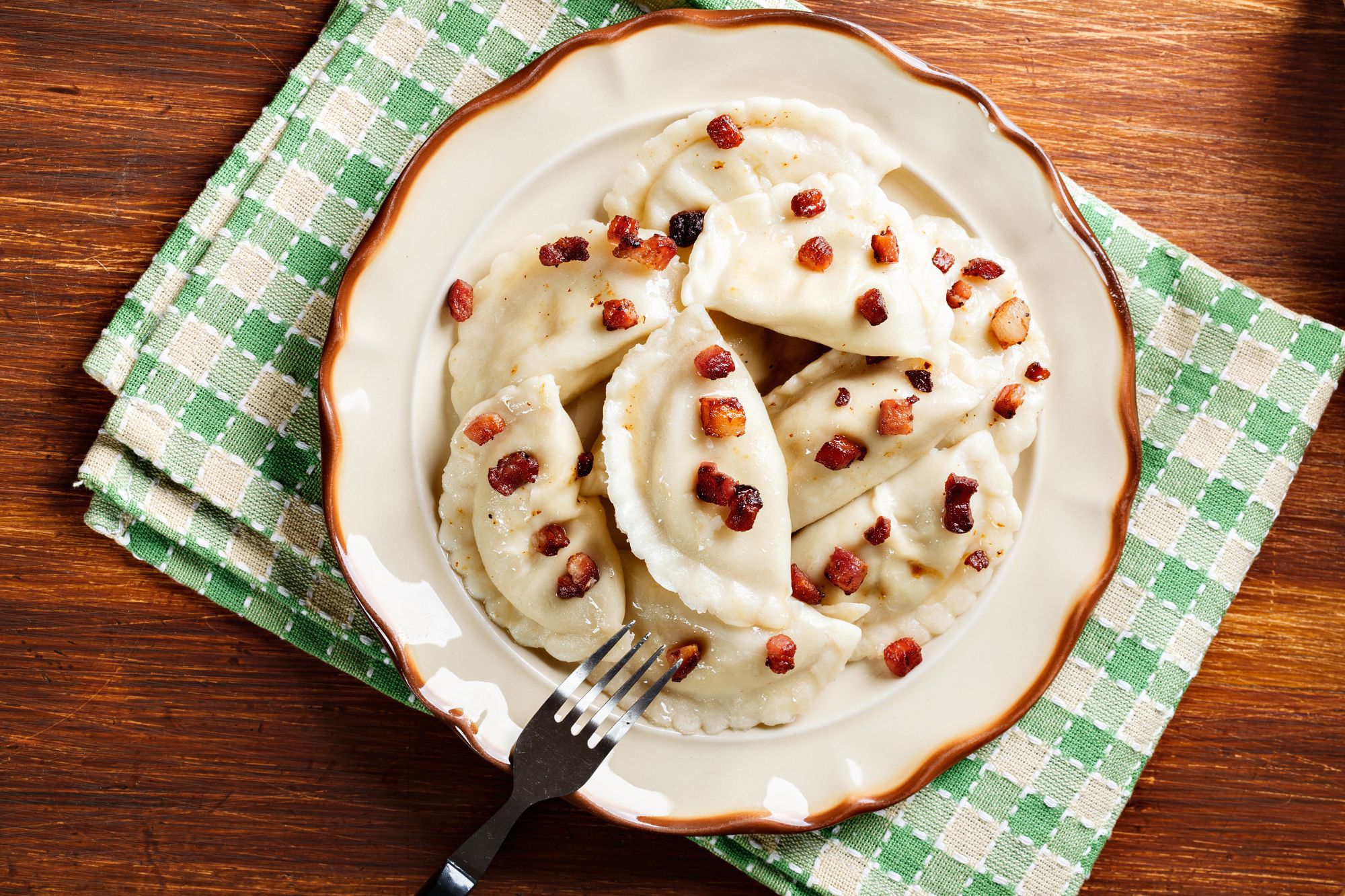 Pierogi z boczkiem na talerzu, z widelcem, na zielonym obrusu w kratkę.