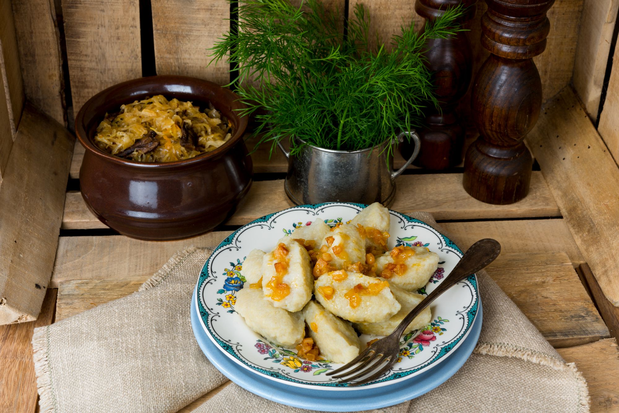 Pierogi z sosem, obok garnka z kapustą i ziołami na drewnianym tle.