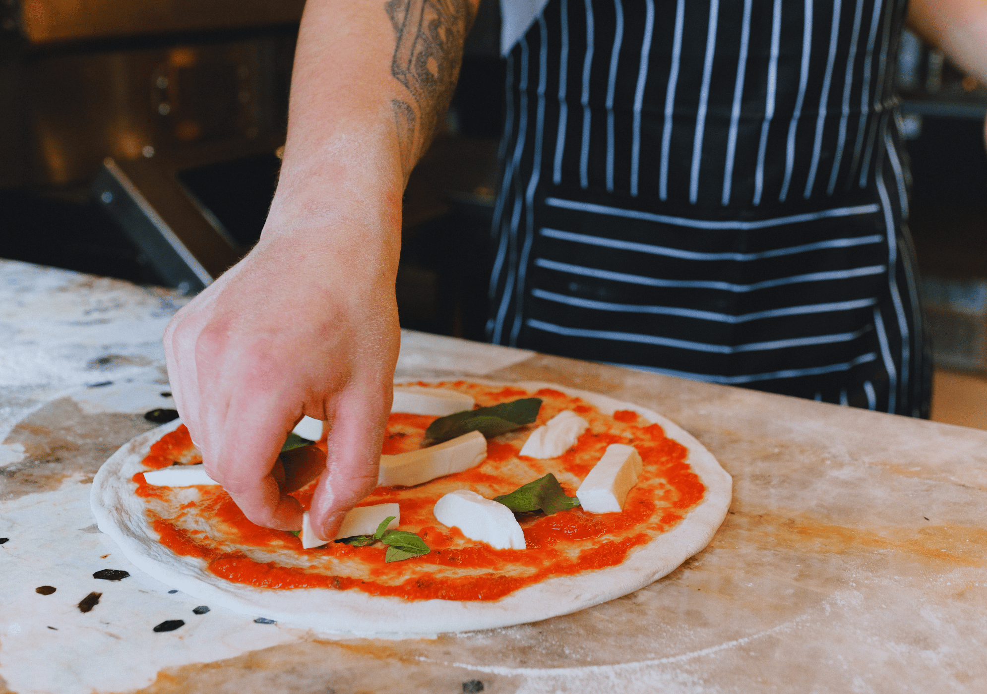Kuchár pripravuje pizzu s paradajkovým základom, syrom a bazalkou na pracovnej doske.