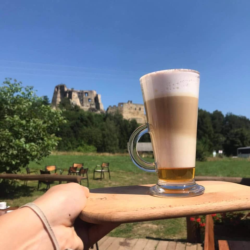 Lódka z kawą latte na drewnianym talerzu z zamkiem w tle, otoczenie zieleni i siedzenia na świeżym powietrzu.