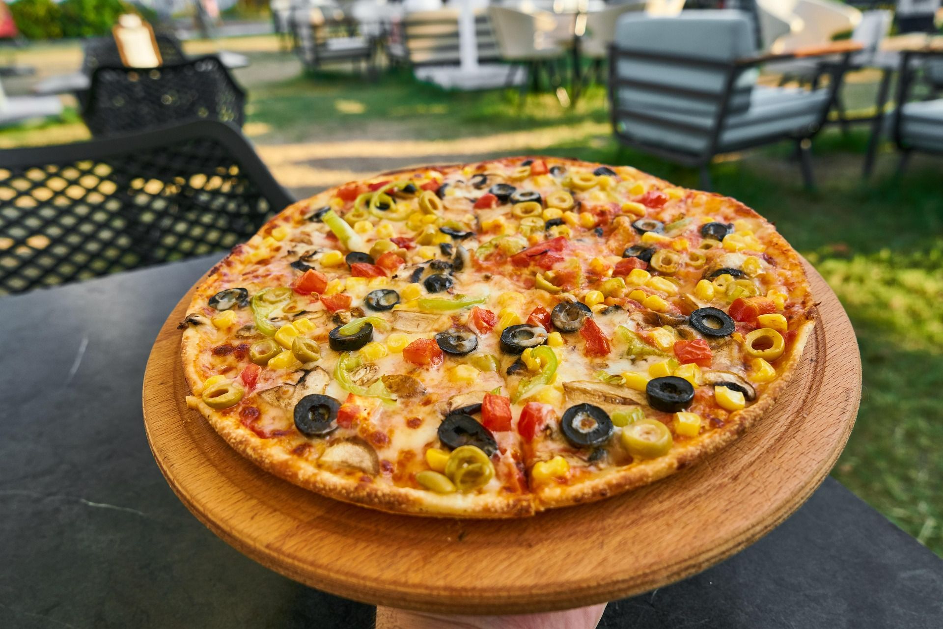 Pizza na drewnianej desce, z serem, pomidorami, oliwkami i papryką, w plenerze w otoczeniu mebli ogrodowych.