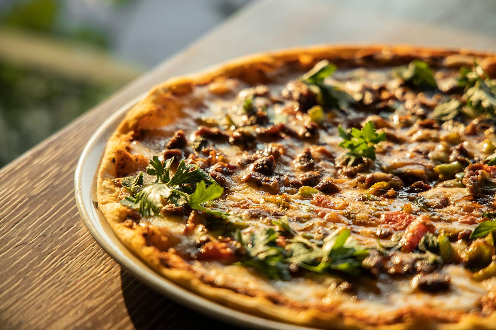 Pizza na talerzu z mięsem i świeżą kolendrą, na drewnianym stole w tle.