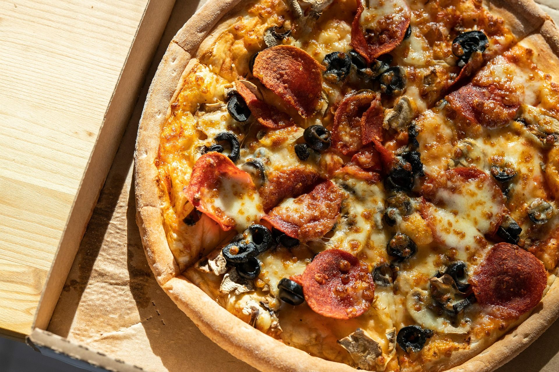 Pizza z pepperoni, oliwkami i serem, podana w kartonowym opakowaniu na drewnianym blacie.