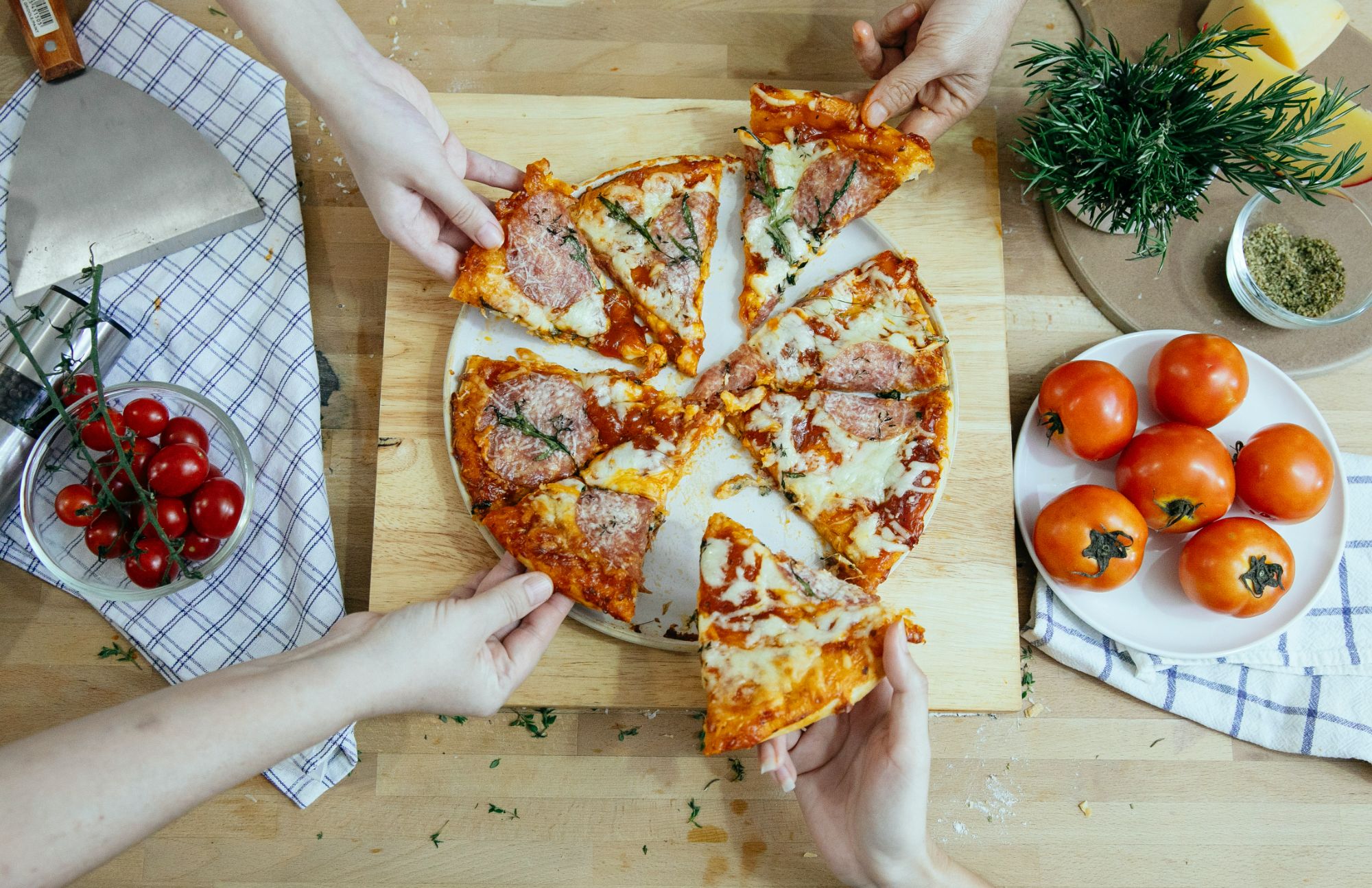 Ręce wyciągające kawałki pizzy z okrągłej deski, obok pomidory i zioła na stole.