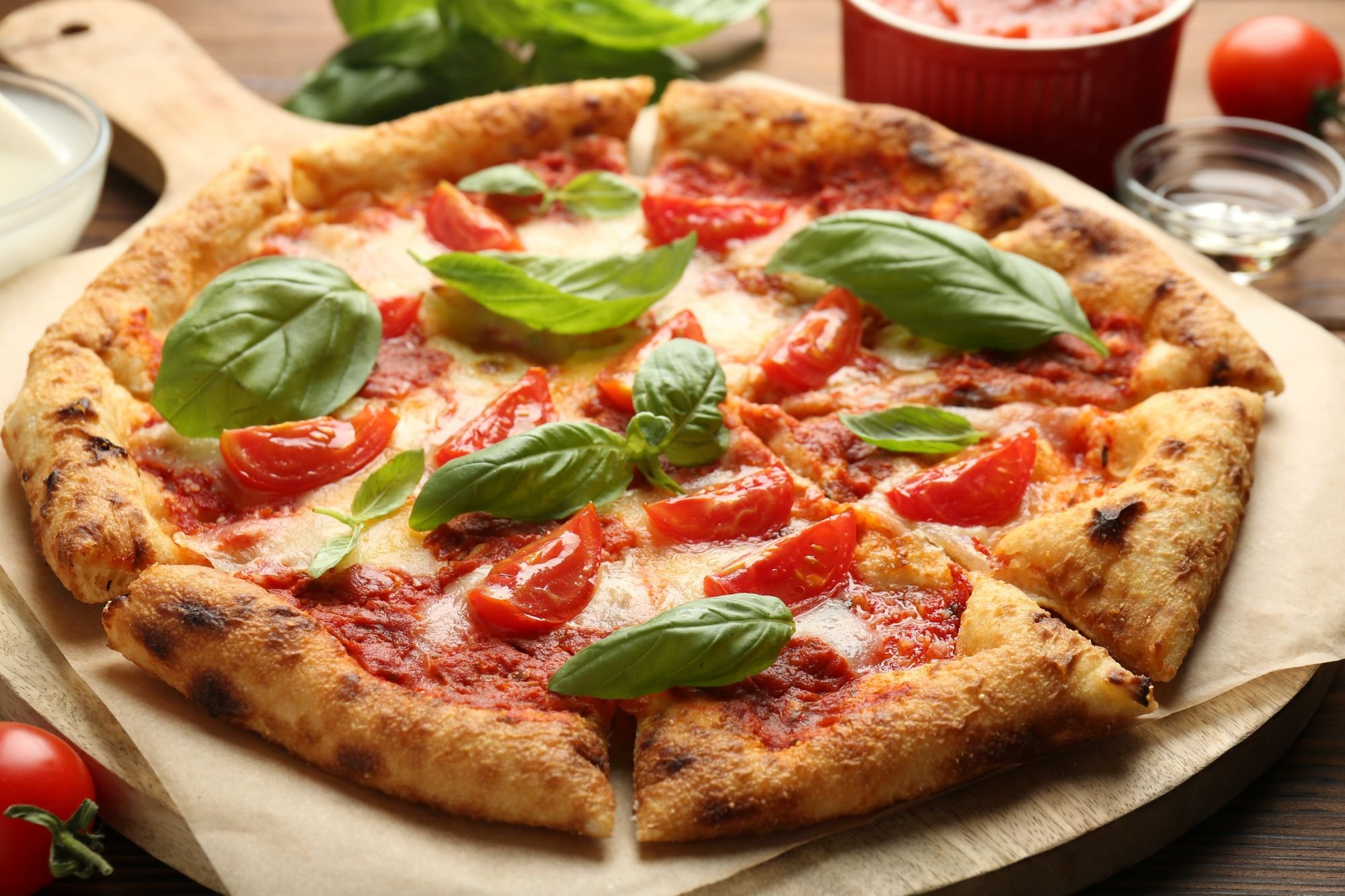 Pyszna pizza z pomidorami, bazylią i serem mozzarella na drewnianej desce, obok sos pomidorowy i składniki.