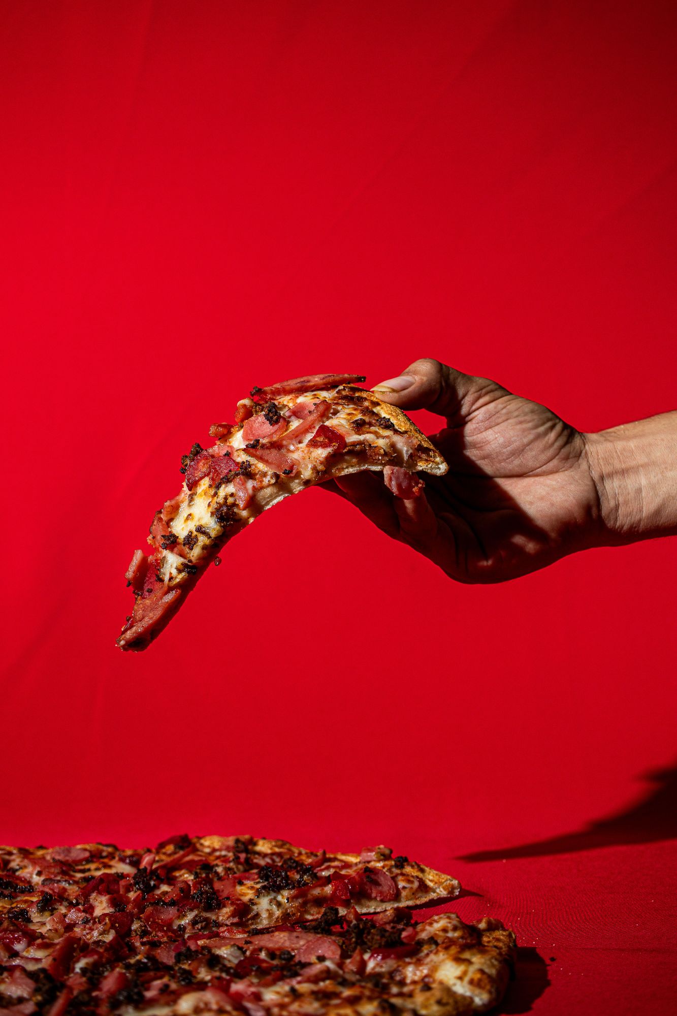 Ręka trzymająca kawałek pizzy z dodatkiem boczku na tle czerwonej tkaniny.
