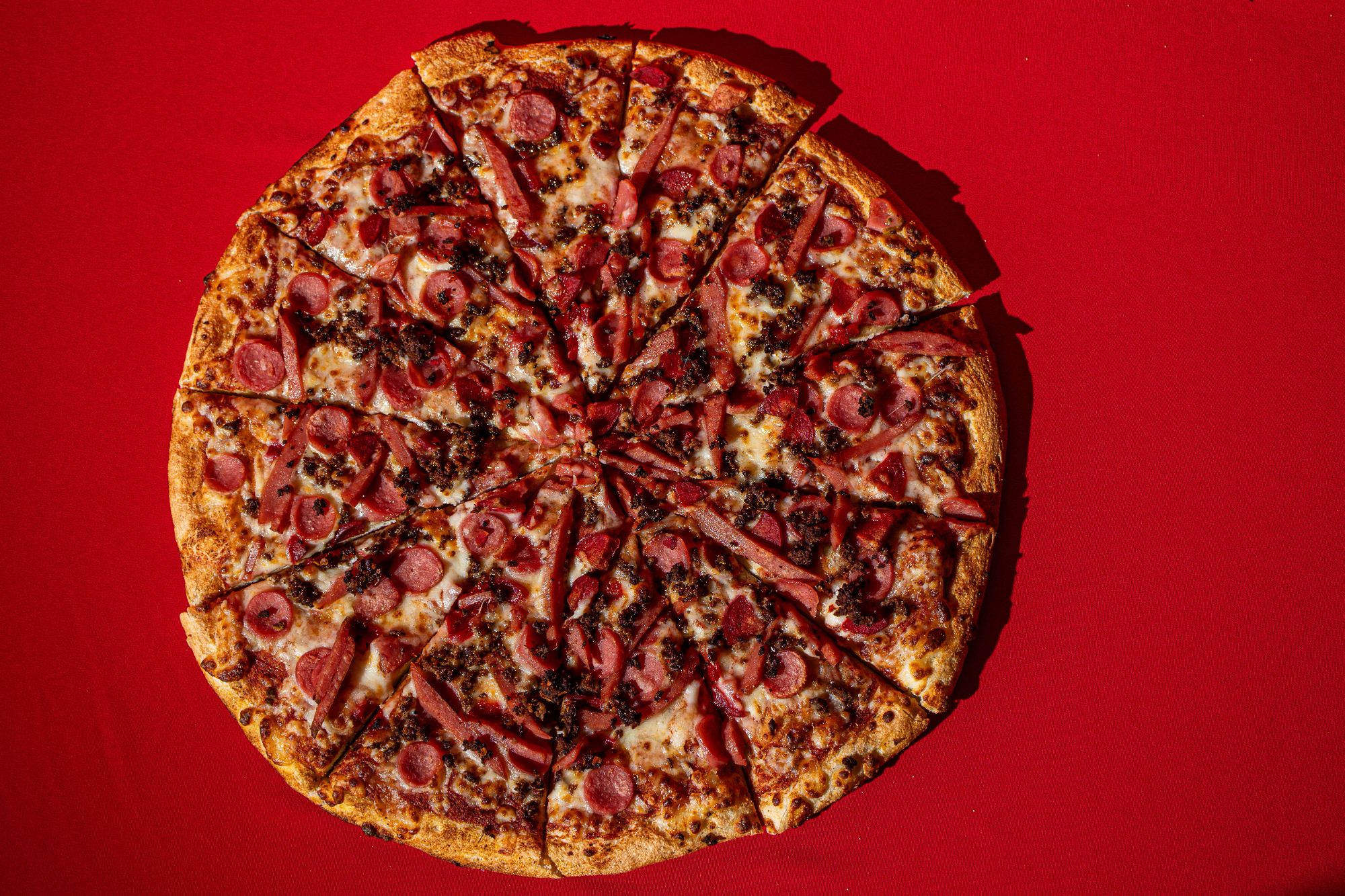 Pizza na czerwonym tle, pokryta plasterkami kiełbasy i przypiekanym serem, pokrojona na kawałki.