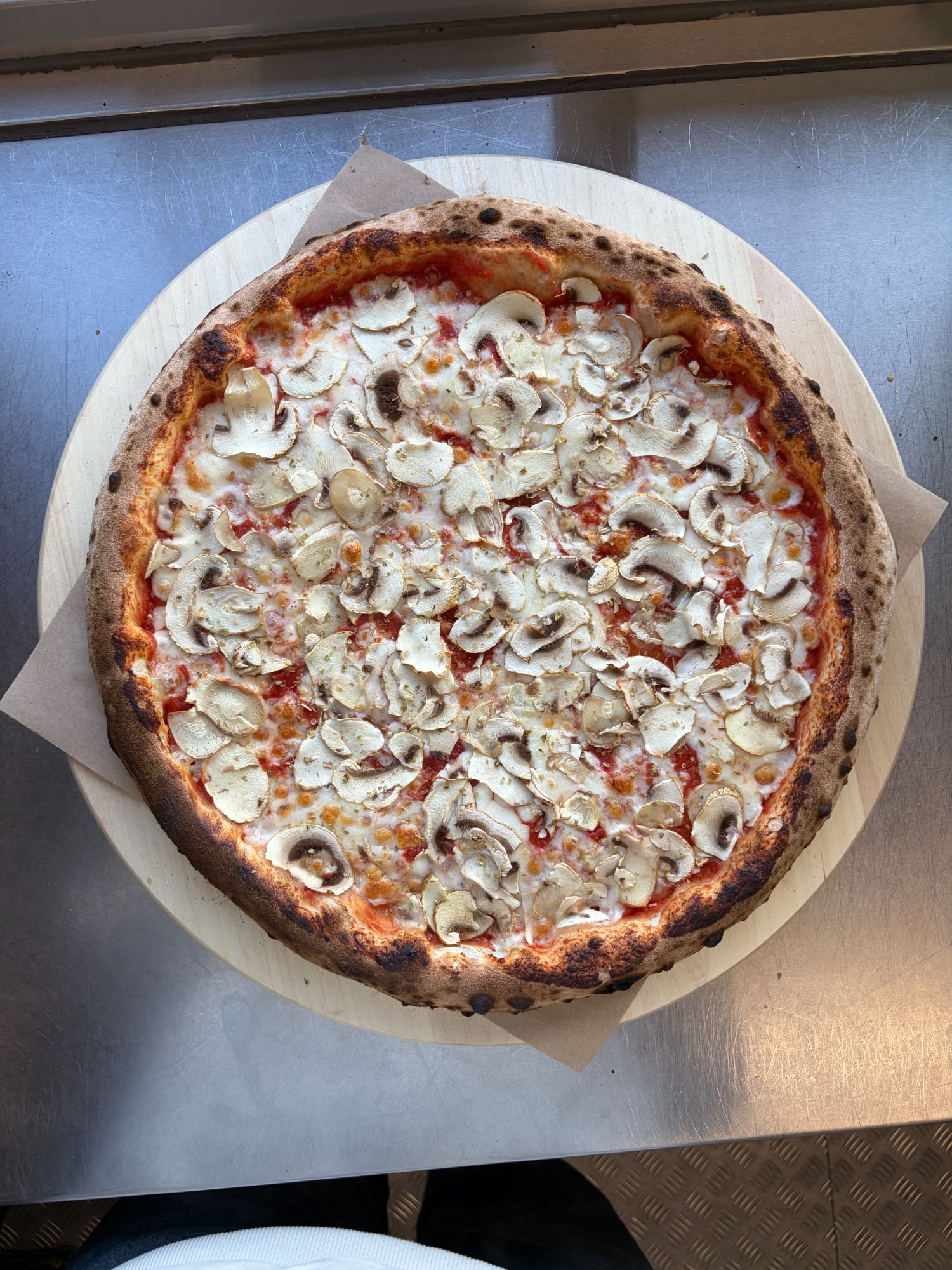 Pizza z serem i pieczarkami na drewnianym talerzu, umieszczona na metalowej powierzchni.