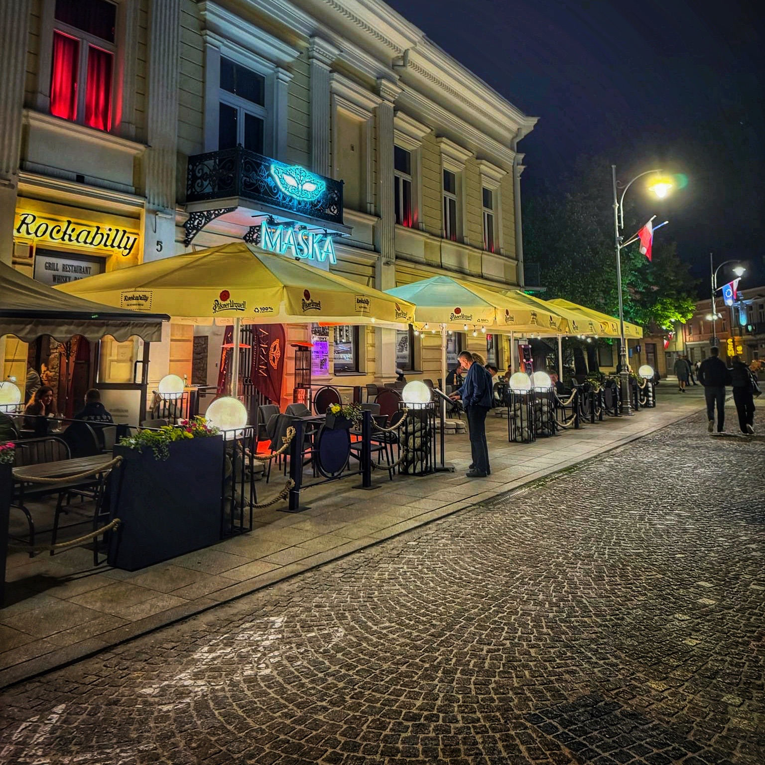 Ulica nocą z oświetlonymi restauracjami i gośćmi siedzącymi na tarasie.