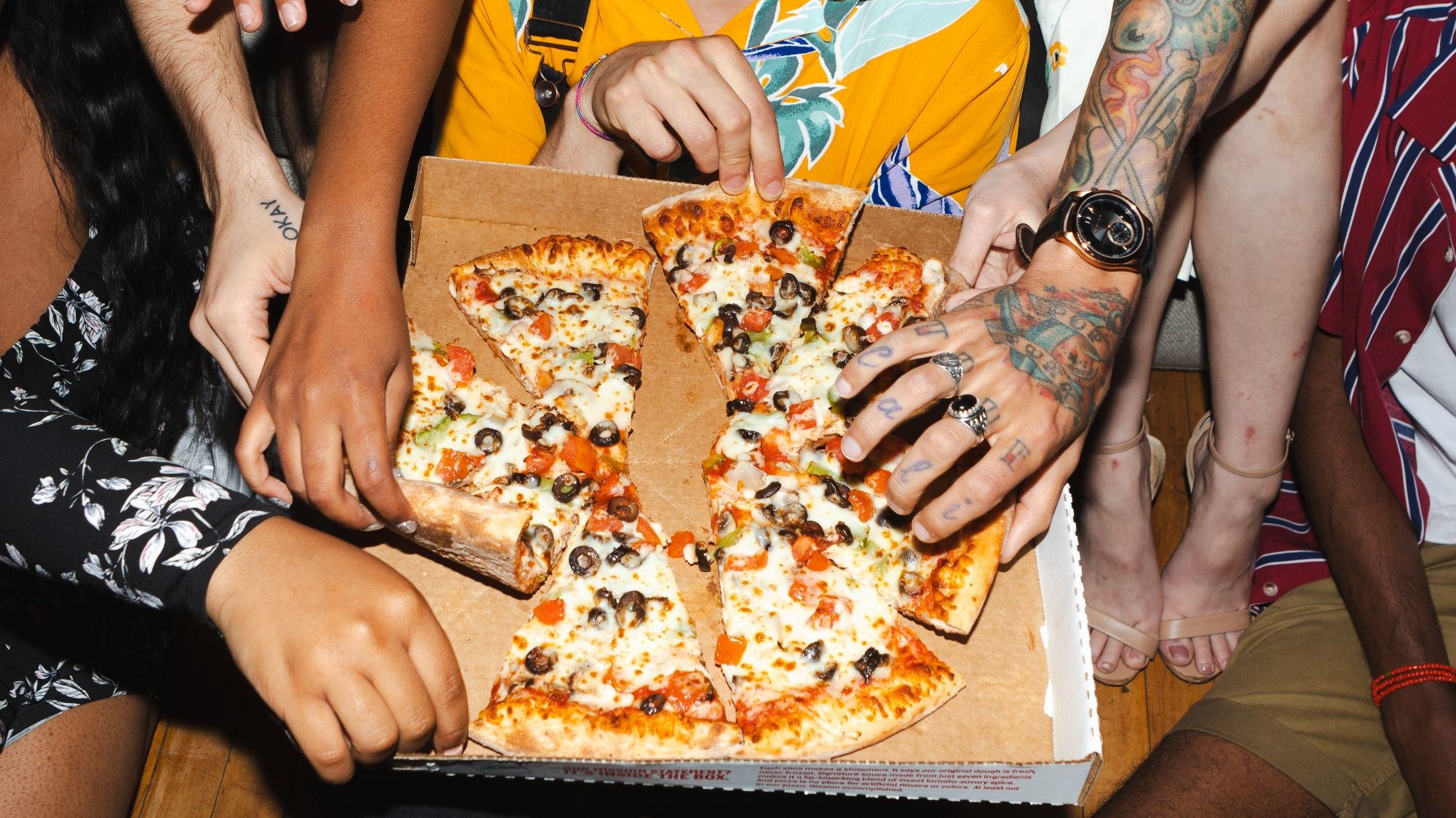 Grupa osób sięga po kawałki pizzy w kartonie, różnorodne składniki, radosna atmosfera.