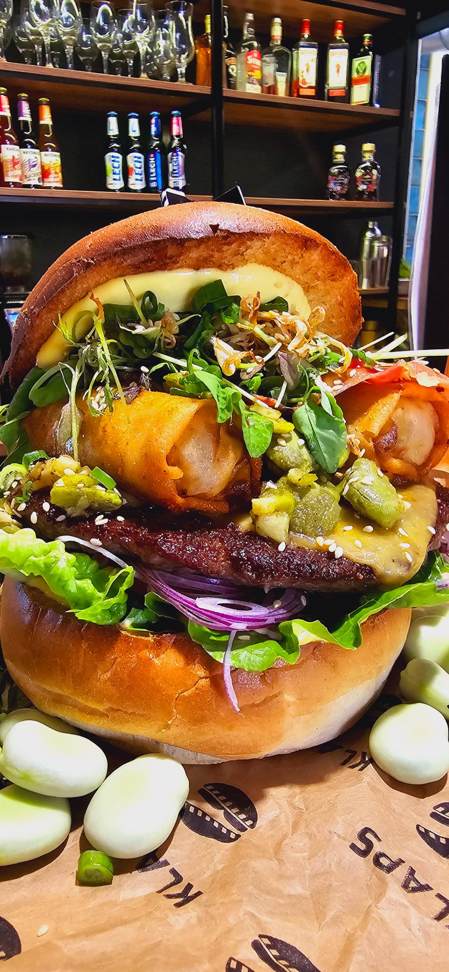 Duży burger z soczystym mięsem, świeżymi warzywami, sosem i dodatkami, w otoczeniu składników na stole.