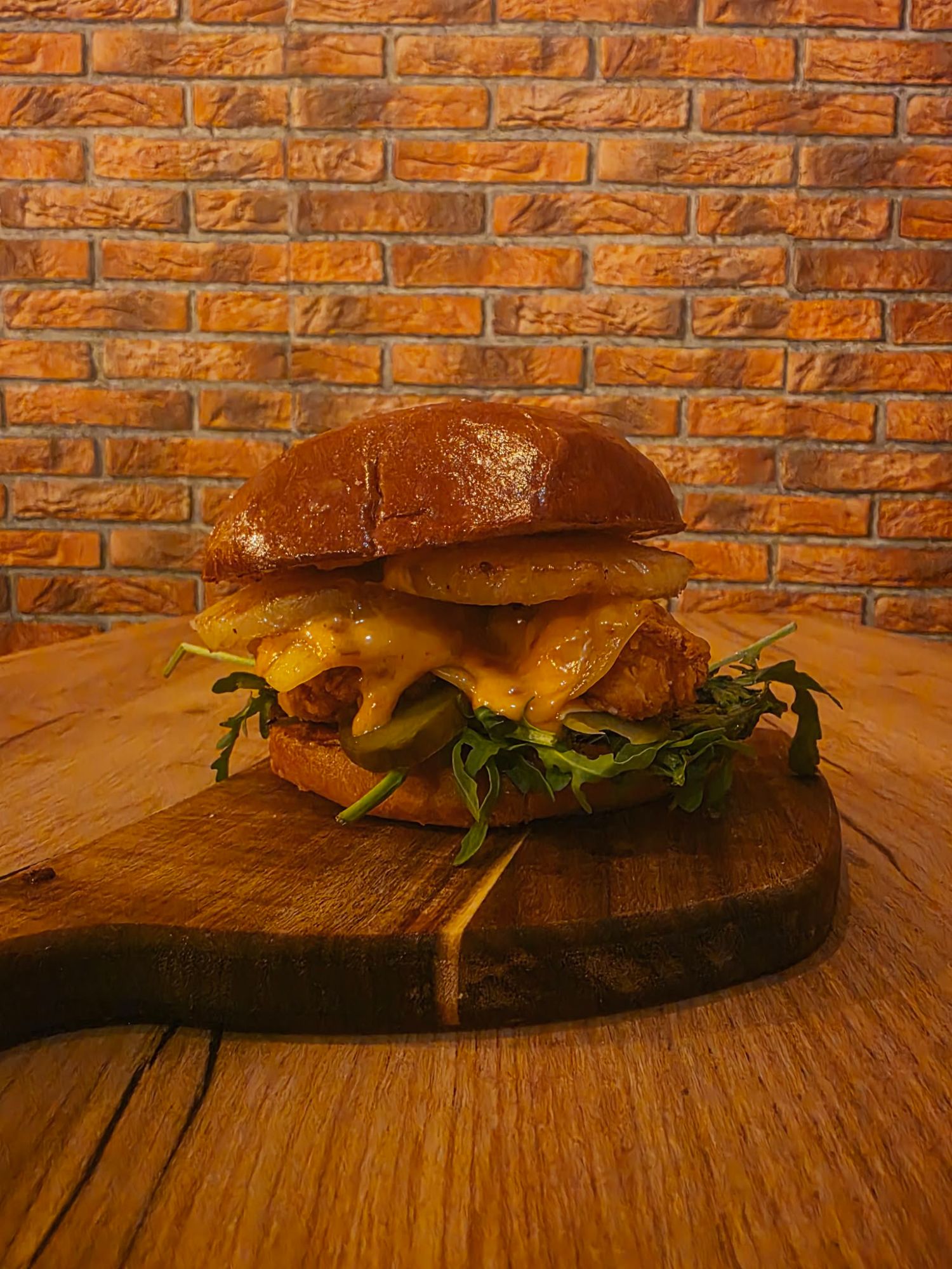 Soczysty burger z kurczakiem, serem cheddar, rukolą i ogórkiem na tle ceglanej ściany.