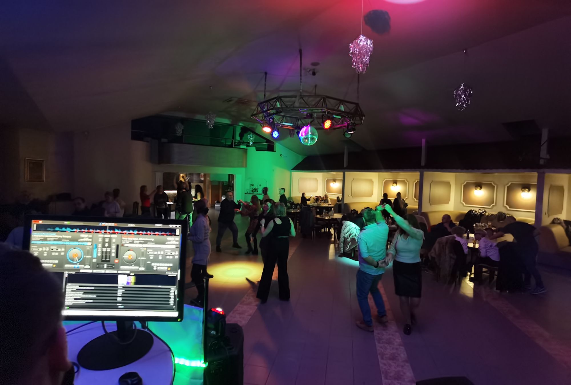 Ludzie tańczą w kolorowym oświetleniu na parkiecie w klubie, przy widocznym stanowisku DJ-a.