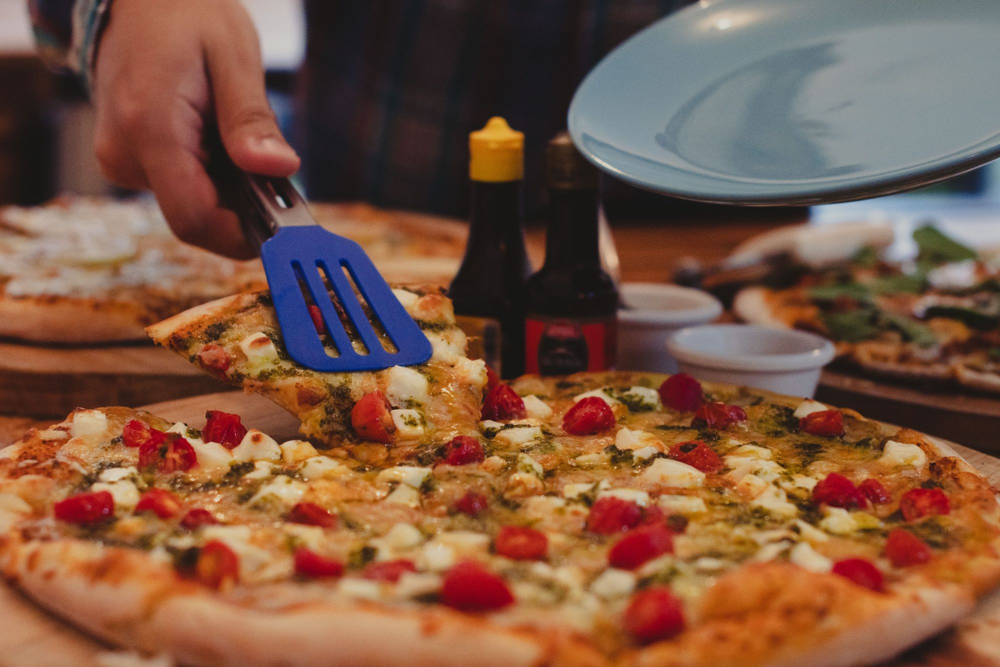 Osoba podaje kawałek pizzy z sosem pesto, pomidorkami i serem, obok butelki z przyprawami.