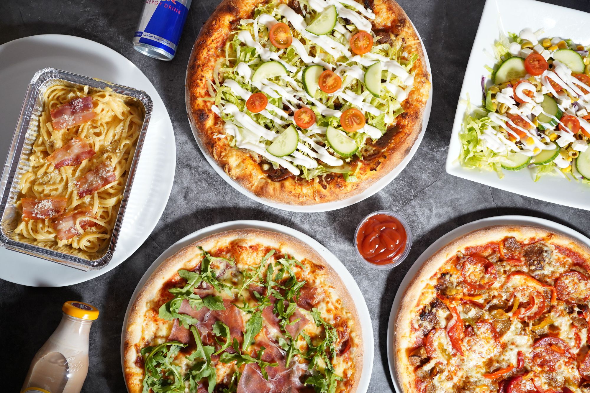 Pizza z różnymi dodatkami, makaron, sałatka oraz napój energetyczny w zestawie na stole.