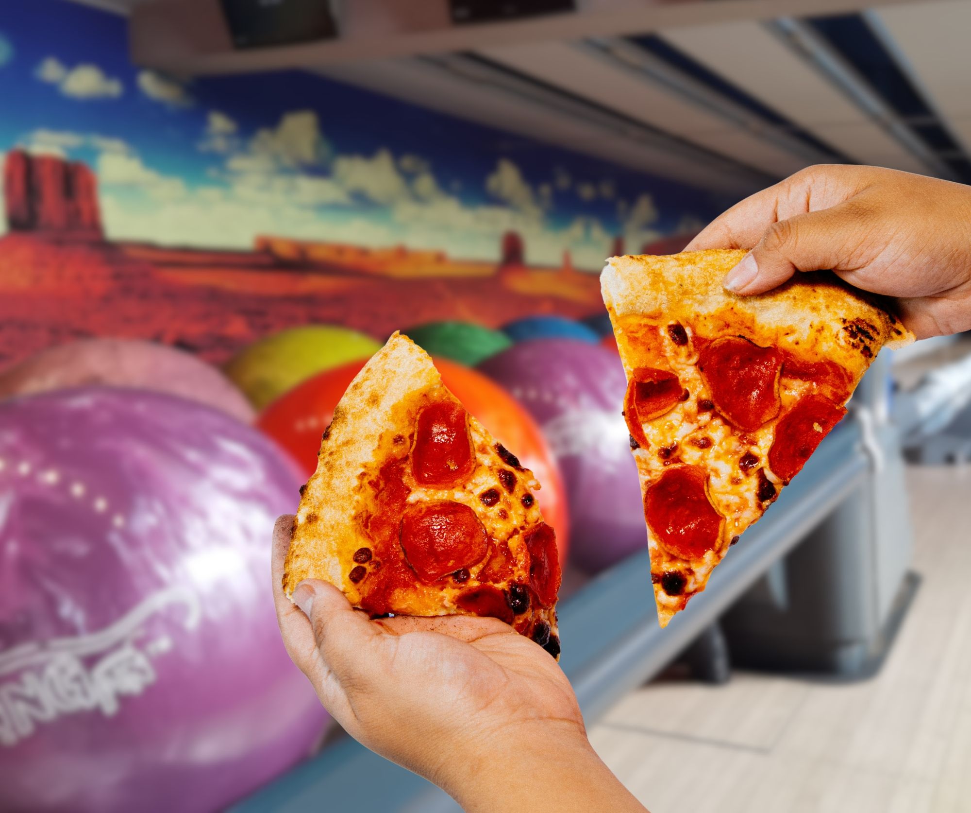 Osoba trzyma dwie kawałki pizzy z pepperoni, w tle kolorowe kule do kręgli i mural krajobrazu.