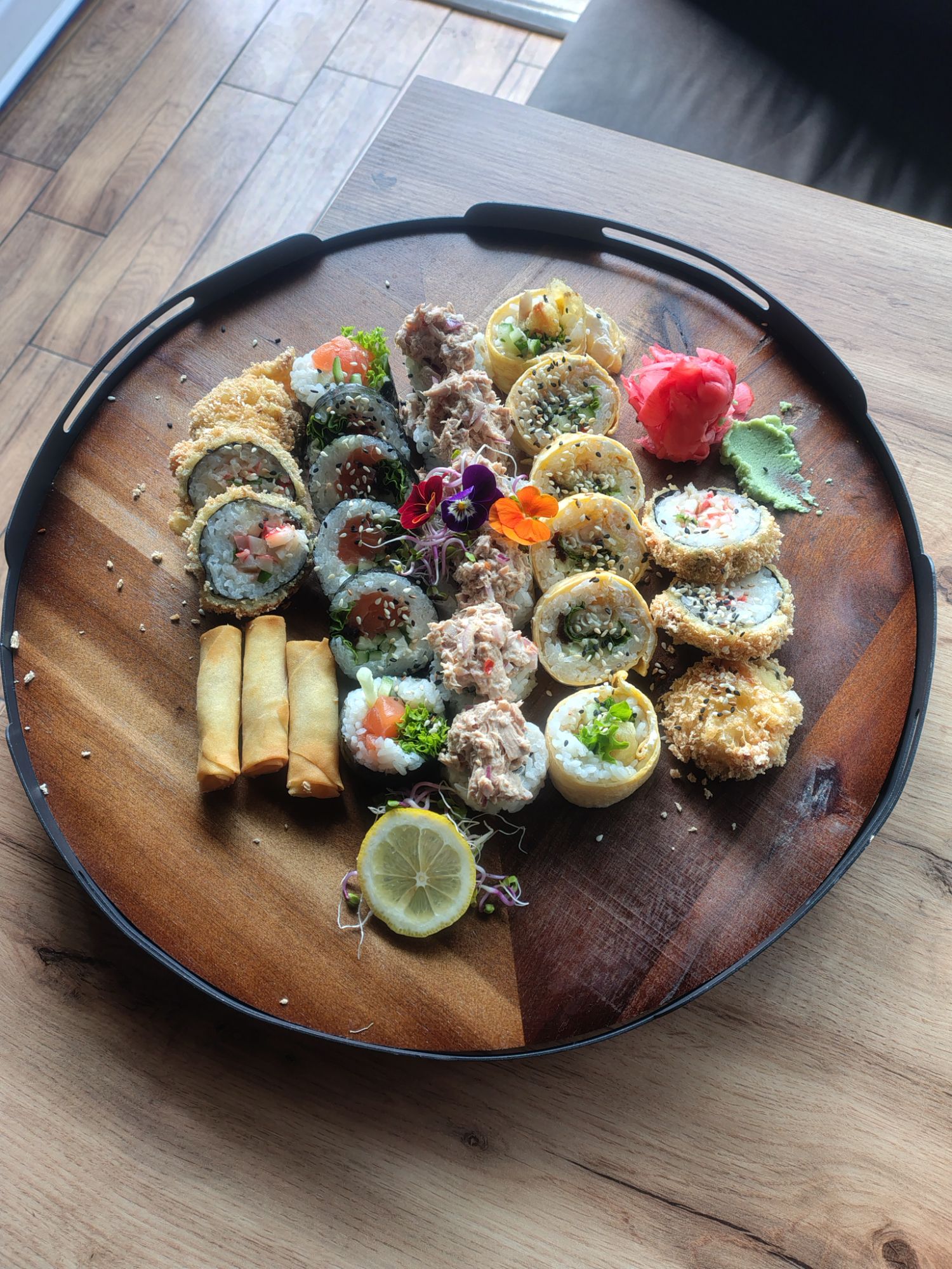 Taca z różnorodnymi sushi, rolkami, krewetkami i wiosennymi rulonikami, udekorowana kwiatami i cytryną.